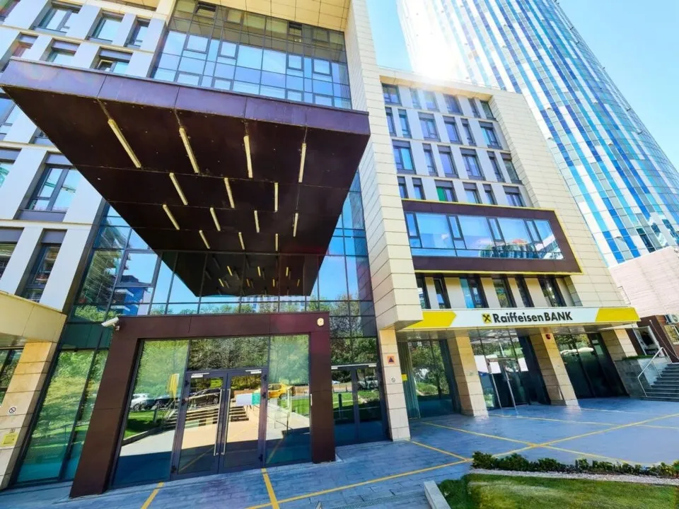 O clădire modernă cu fațadă de sticlă și panouri de piatră prezintă o intrare cu un acoperiș în consolă și un panou galben cu text.