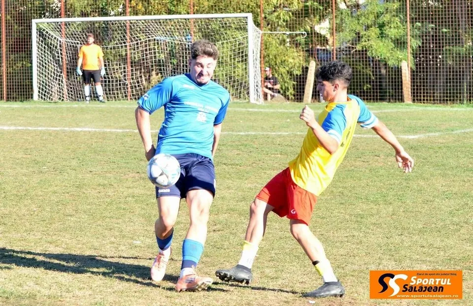 Un jucător de fotbal într-un tricou albastru conduce mingea pe un teren de iarbă, fiind urmărit de un adversar în tricou galben și pantaloni roșii.