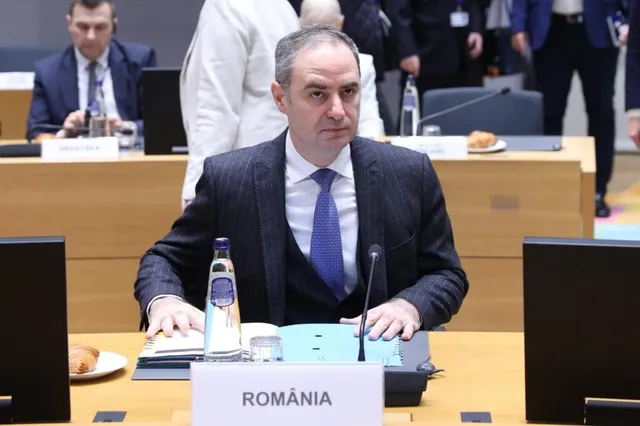 Un bărbat într-un costum închis și cravată albastră stă la o masă de conferință lângă o sticlă de apă și un monitor.