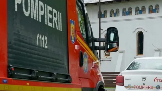 Un camion de pompieri roșu și o mașină albă cu inscripții sunt parcate în fața unei clădiri albe cu ferestre arcuite și picturi murale.