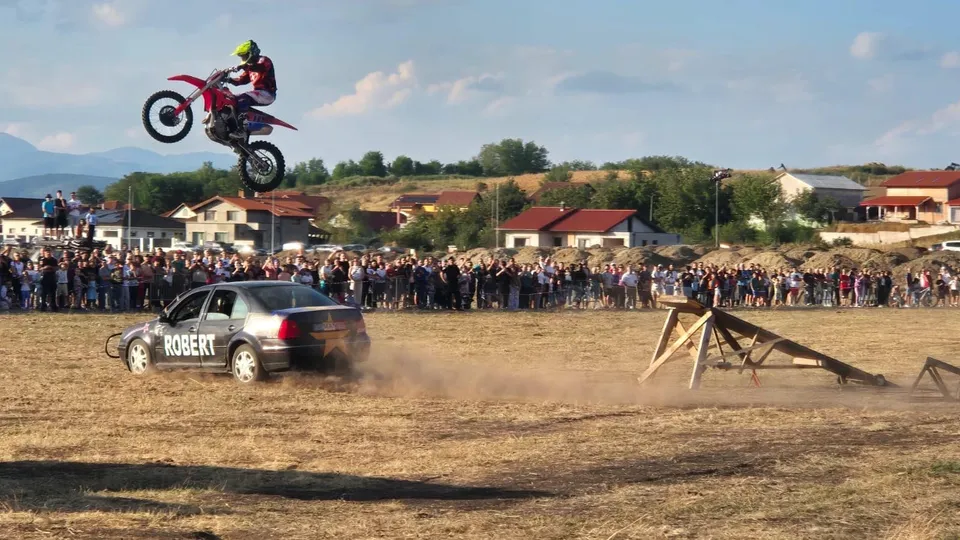 Un motociclist cu cască galbenă execută un salt cu o motocicletă roșie deasupra unei mașini gri într-un câmp deschis în fața unei mulțimi.