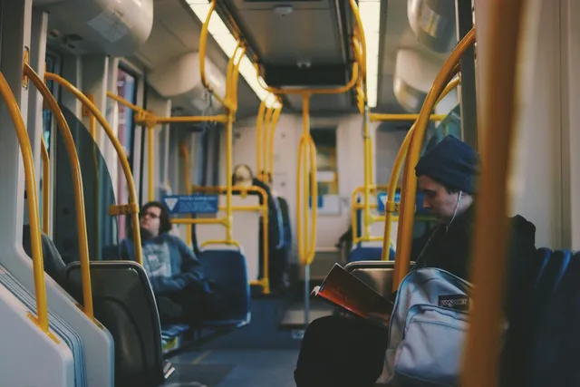 Un bărbat cu căciulă și un rucsac stă pe un scaun într-un autobuz lângă barele de prindere galbene.