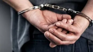 Mâinile unui bărbat sunt prinse în cătușe în spatele spatelui, fiind purtate de o persoană îmbrăcată într-un tricou gri și pantaloni închiși la culoare.