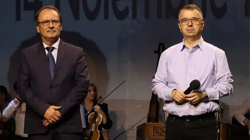 Un bărbat în costum închis și unul în cămașă deschisă stau în picioare în fața unui fundal întunecat, lângă un violonist.