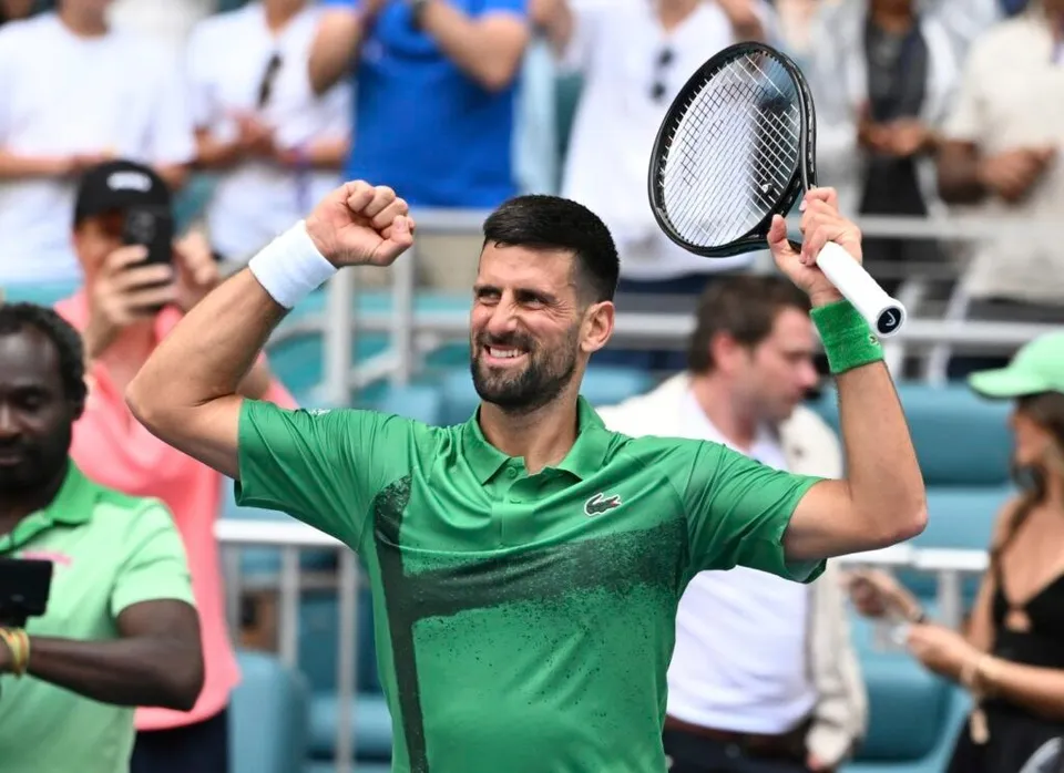 Un bărbat cu tricou verde și brățări albe și verzi ridică brațele și un rachete de tenis pe un teren de sport.