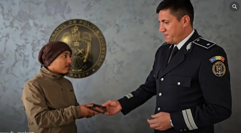 Un bărbat cu căciulă maro și o jachetă maro primește un obiect mic de la un polițist în uniformă în fața unui emblema circulară pe perete.
