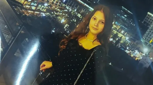 O fată cu păr lung, închis la culoare, poartă o rochie neagră cu buline albe și un colier auriu, pe fundalul unui oraș iluminat noaptea.