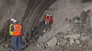 Trei persoane purtând căști de protecție și veste reflectorizante stau lângă o structură de beton și un perete de pământ într-un șantier.