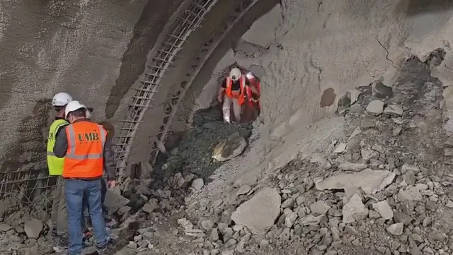 Trei persoane purtând căști de protecție și veste reflectorizante stau lângă o structură de beton și un perete de pământ într-un șantier.