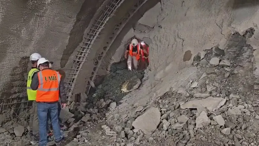 Trei persoane purtând căști de protecție și veste reflectorizante stau lângă o structură de beton și un perete de pământ într-un șantier.