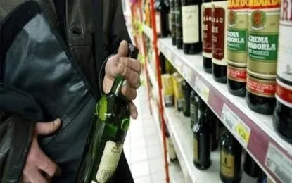 Un bărbat ține o sticlă de vin verde lângă un raft de supermarket plin cu diverse tipuri de băuturi alcoolice.