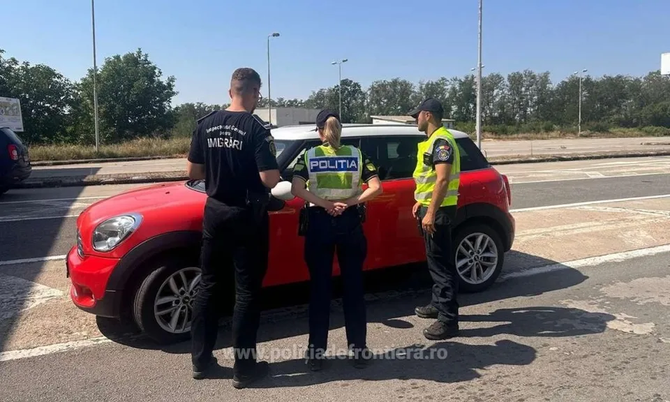 Trei persoane în uniforme stau lângă o mașină roșie pe un drum asfaltat sub un cer senin.