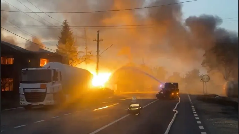 Un camion alb și un vehicul negru circulă pe un drum asfaltat lângă un incendiu cu flăcări și fum dens.