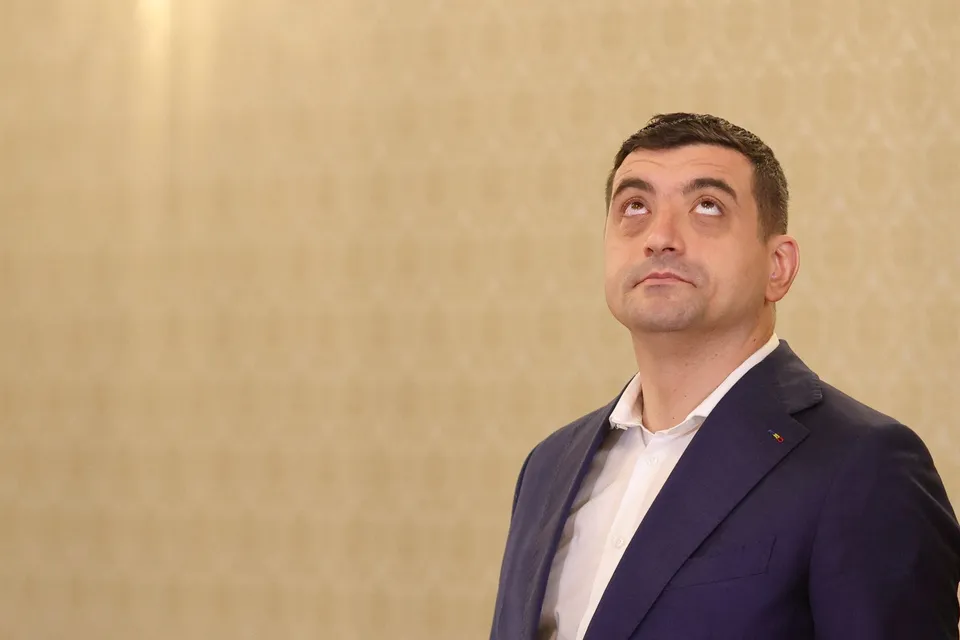 Un bărbat cu părul scurt și costum albastru privește în sus în fața unui perete cu tapet decorativ bej.