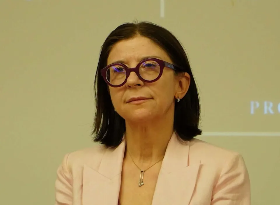 O femeie cu părul negru, purtând ochelari cu rame roșii și un sacou roz deschis, privește spre camera foto.