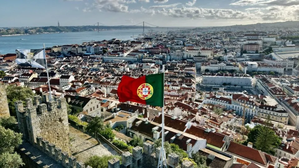 Steagul Portugaliei este agitat pe un stârf în prim-plan, deasupra unei panorame urbane cu clădiri cu acoperișuri roșii și un fluviu în fundal.