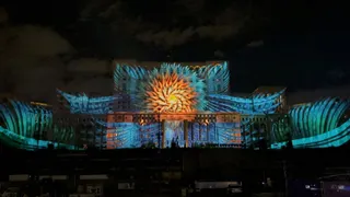 O clădire mare este iluminată de proiecții colorate cu modele geometrice și forme florale pe fațadă sub un cer nocturn.