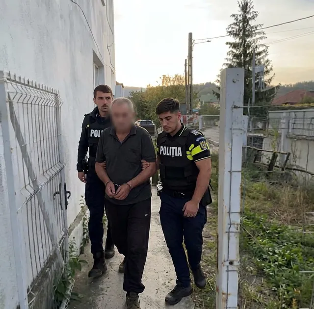 Doi polițiști în uniformă conduc un bărbat cu mâinile în față, legat cu cătușe, pe un drum îngust lângă un zid alb.