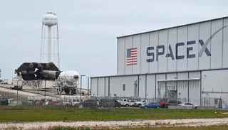 Un hangar mare cu inscripția SpaceX, un turn de apă alb și o rachetă deasupra unei platforme de lansare sub un cer înnorat.