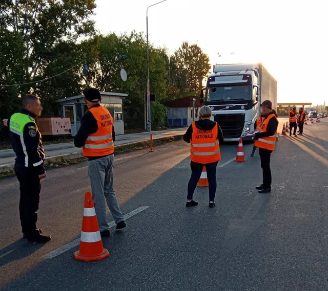 Patru persoane purtând veste reflectorizante portocalii stau pe un drum asfaltat lângă un camion alb și conuri de trafic portocalii.