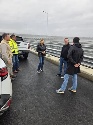 Cinci persoane stau pe un drum asfaltat lângă un pickup alb și o barieră metalică de protecție sub un cer înnorat.