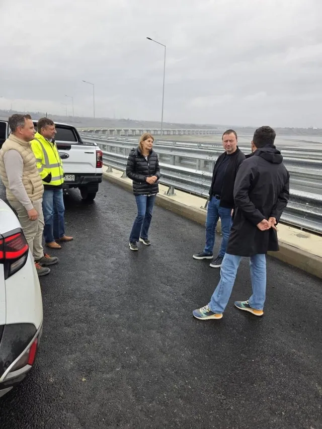 Cinci persoane stau pe un drum asfaltat lângă un pickup alb și o barieră metalică de protecție sub un cer înnorat.