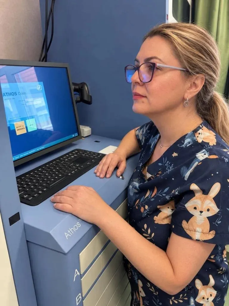 O femeie cu ochelari și uniformă albastră cu modele de animale stă la un birou cu monitor și tastatură.