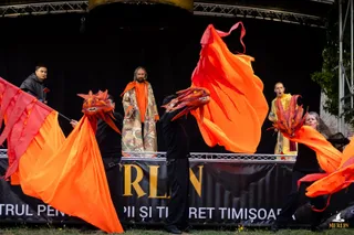 Actori pe o scenă de exterior manipulează dragoni din material textil portocaliu și roșu sub un decor cu un personaj într-o robă lungă.