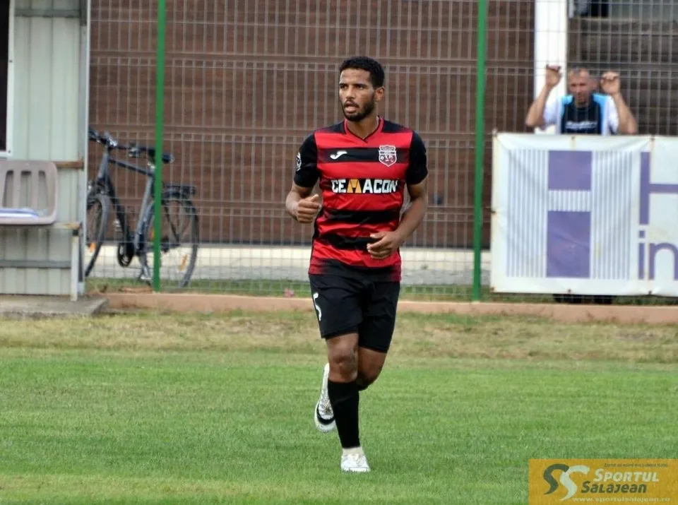 Un bărbat cu tricou roșu și negru cu dungi orizontale aleargă pe un teren de fotbal cu iarbă verde.