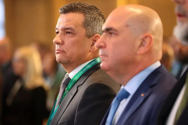 Doi bărbați în costume și cravate stau în picioare într-o încăpere cu fundal desfocat.