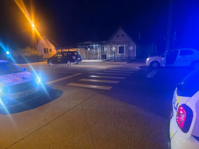 Două mașini de poliție și un autovehicul negru sunt parcate pe un drum lângă un zebru, sub lumina unui stâlp de iluminat nocturn.
