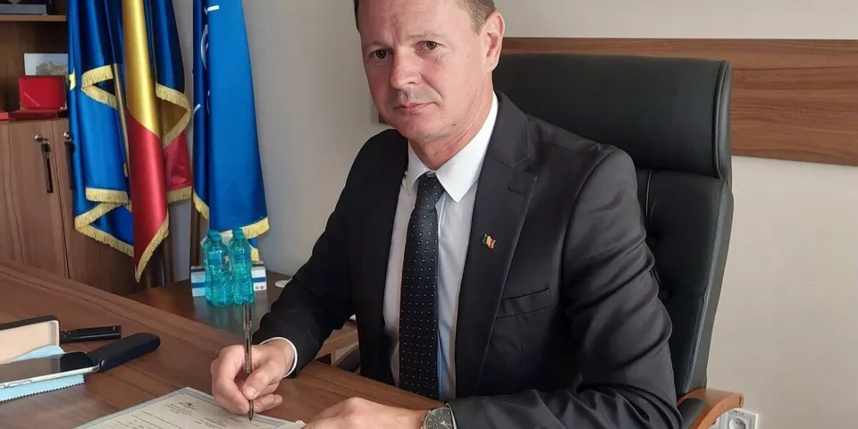 Un bărbat într-un costum închis și cravată stă la un birou și ține un pix deasupra unui document.