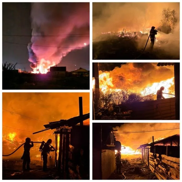 Siluete de pompieri luptă cu flăcările și fumul dens în timpul unui incendiu nocturn lângă structuri de lemn.