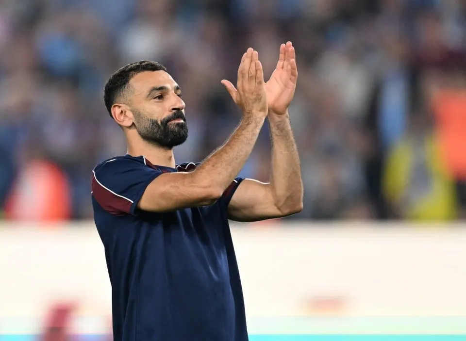 Un bărbat cu barbă și tricou închis la culoare aplaudă cu ambele mâini în timp ce privește spre lateral.