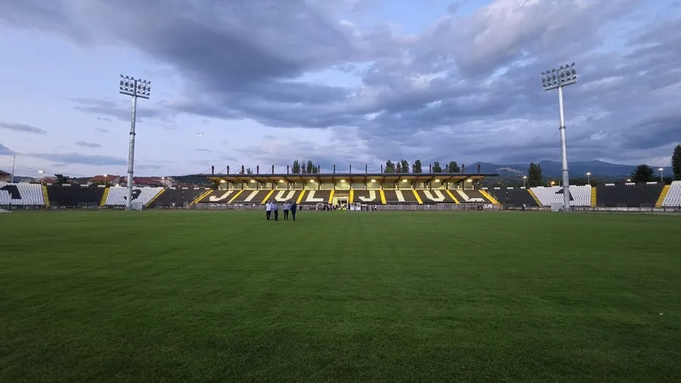 Un teren de fotbal cu iarbă verde este în fața unor tribune cu scaune galbene și negre sub un cer înnorat.