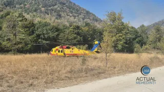 Un elicopter galben și roșu este aterizat pe un câmp de iarbă uscată lângă o pădure de copaci verzi și un deal împădurit.