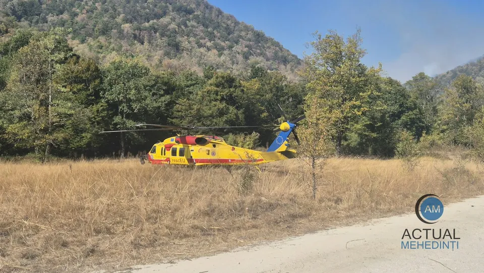 Un elicopter galben și roșu este aterizat pe un câmp de iarbă uscată lângă o pădure de copaci verzi și un deal împădurit.