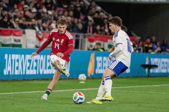 Un jucător în echipament roșu și alb driblează un fotbal pe un teren de fotbal verde în fața unui alt jucător în echipament alb și albastru.