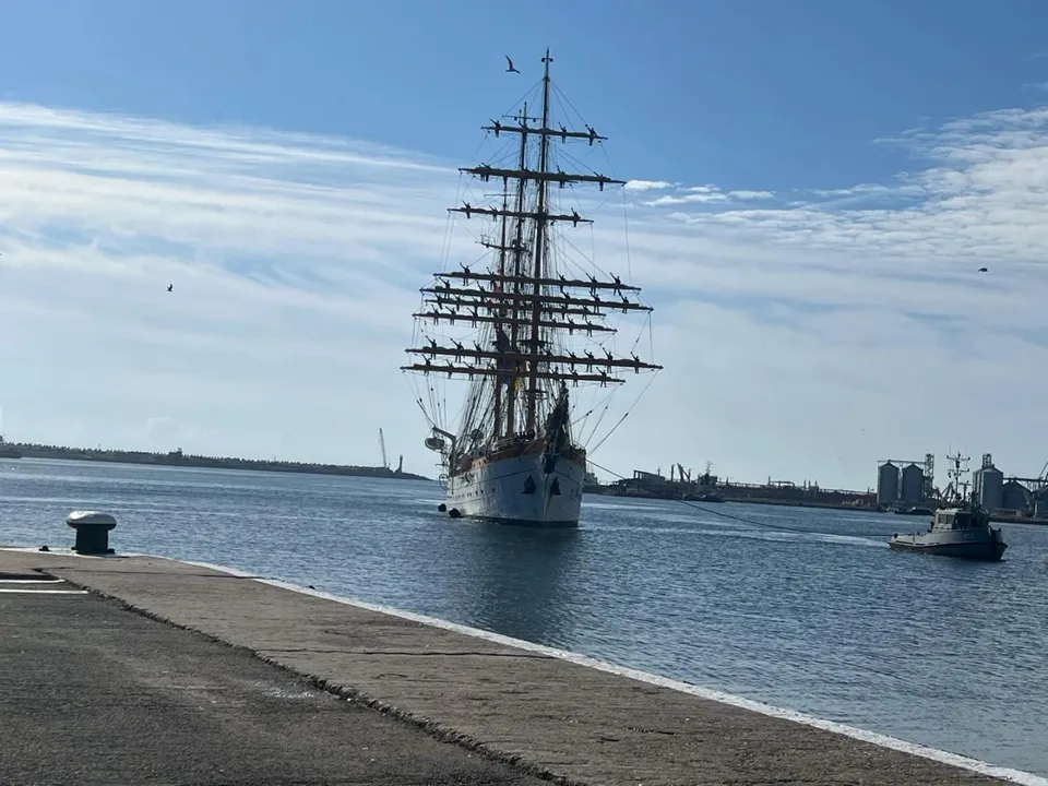 O navă cu trei catarge și vele strânse, cu un remorcher în dreapta, navighează pe un canal, cu un chei în prim-plan și siluete de clădiri portuare în fundal.
