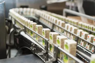 Linii de producție transportă cutii de carton mici, albe, cu imprimeuri verzi, pe o bandă de rulare metalică într-un mediu industrial.