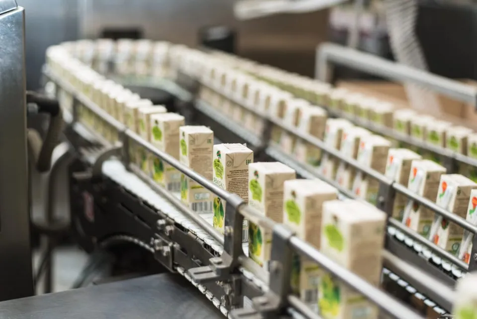 Linii de producție transportă cutii de carton mici, albe, cu imprimeuri verzi, pe o bandă de rulare metalică într-un mediu industrial.