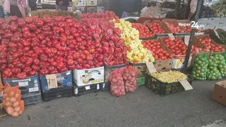 Diverse cantități de roșii, mere și alte fructe sunt expuse în cutii de plastic și pe pământ într-o piață în aer liber.