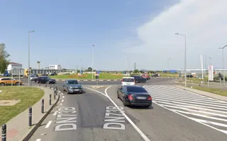 Mașini circulă pe o intersecție asfaltată cu marcaje rutiere albe, sub un cer senin, înconjurată de stâlpi de iluminat și vegetație discretă.