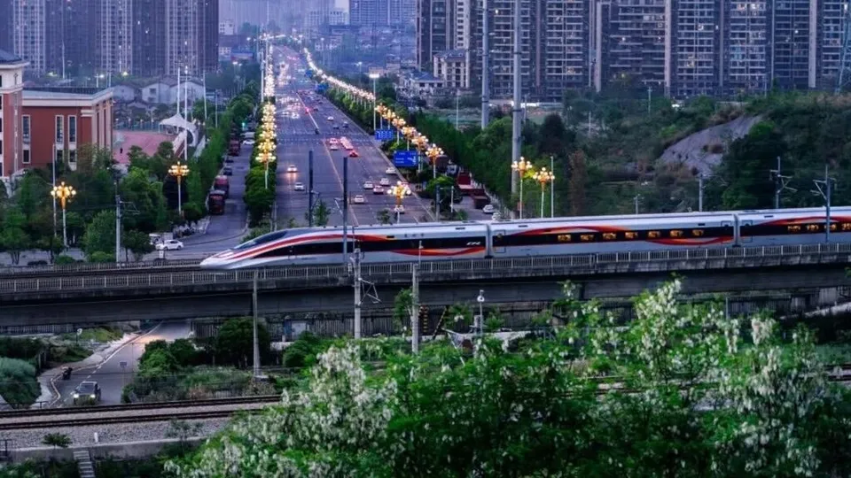 Un tren de mare viteză cu culori alb, roșu și negru circulă pe un viaduct ridicat într-un peisaj urban cu clădiri și drumuri luminat.