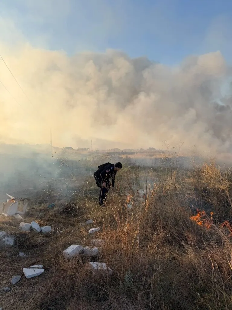 O persoană în haine închise stă aplecată într-un câmp cu iarbă uscată, lângă un foc mic și resturi albe de materiale.