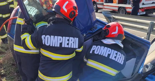 Două persoane purtând uniforme de pompieri cu benzi reflectorizante galben-verzi lucrează lângă un vehicul albastru pe o stradă.