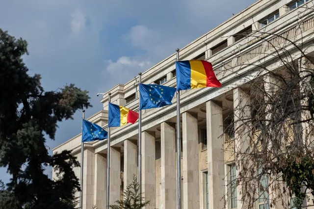 Trei steaguri ale Uniunii Europene și un steag tricolor sunt arborate pe stâlpi în fața unei clădiri cu coloane înalte sub un cer înnorat.