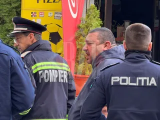 Trei bărbați în uniforme de serviciu, unul cu inscripția POMPIER și unul cu POLITIA, stau în fața unei clădiri cu un panou galben.