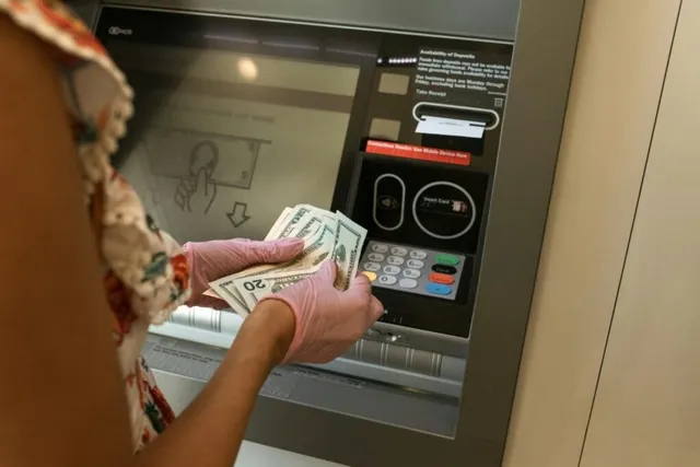O persoană cu mănuși roz ține mai multe bancnote de douăzeci de dolari în fața unui ecran de bancomat.