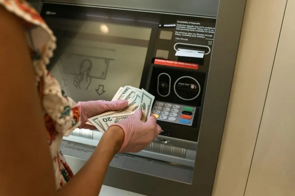 O persoană cu mănuși roz ține mai multe bancnote de douăzeci de dolari în fața unui ecran de bancomat.
